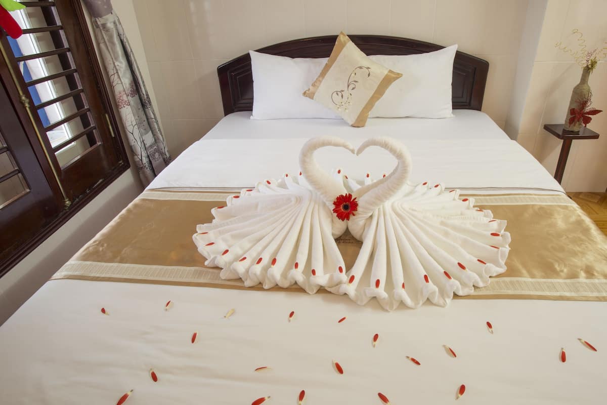 A neatly made bed with a heart-shaped towel arrangement and scattered rose petals.