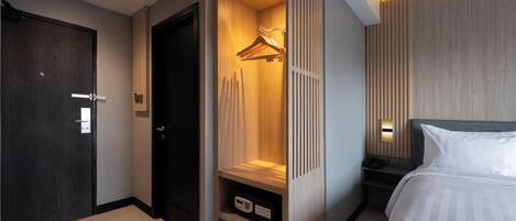 A modern hotel room with a dark wooden door, a wardrobe with a hanging rack, a bedside table with a lamp, and a bed with white bedding.