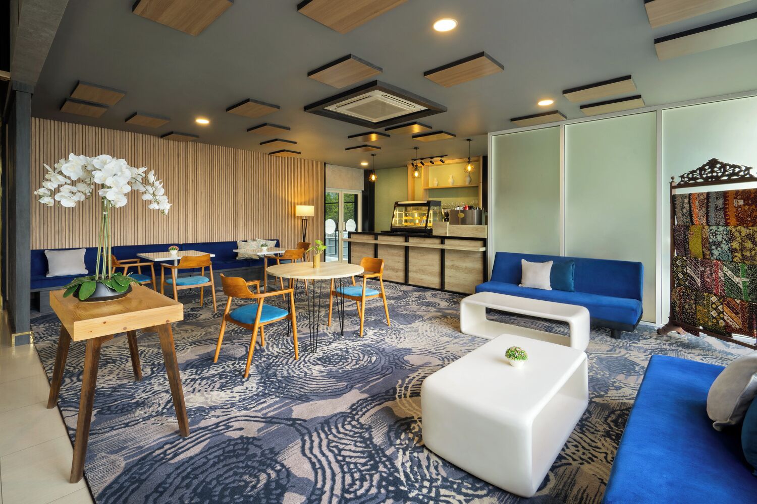 A modern hotel lobby with a blue sofa, a round dining table with chairs, a coffee bar, and a patterned carpet.