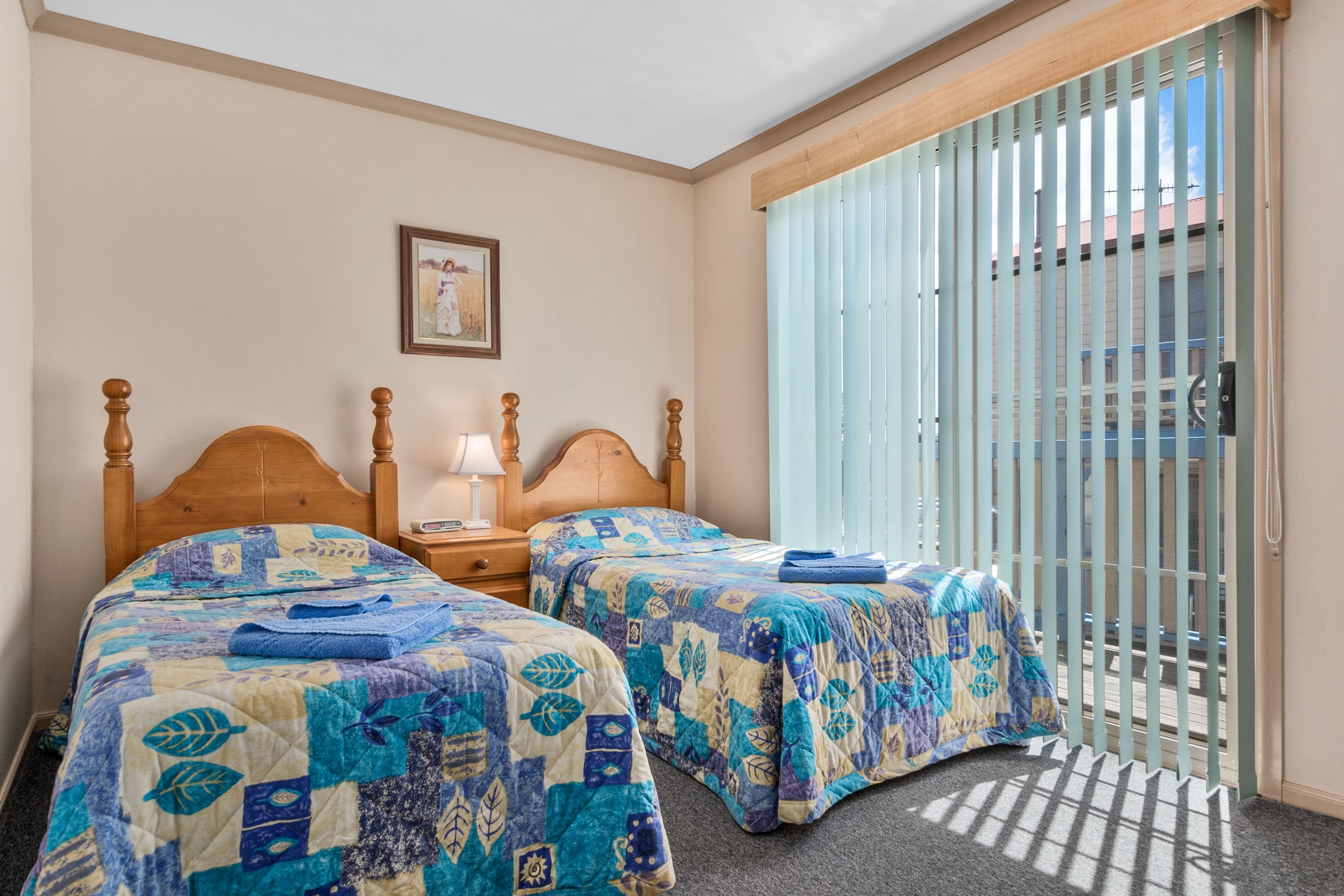 A bedroom with two beds, a nightstand, a lamp, and a window with blinds.