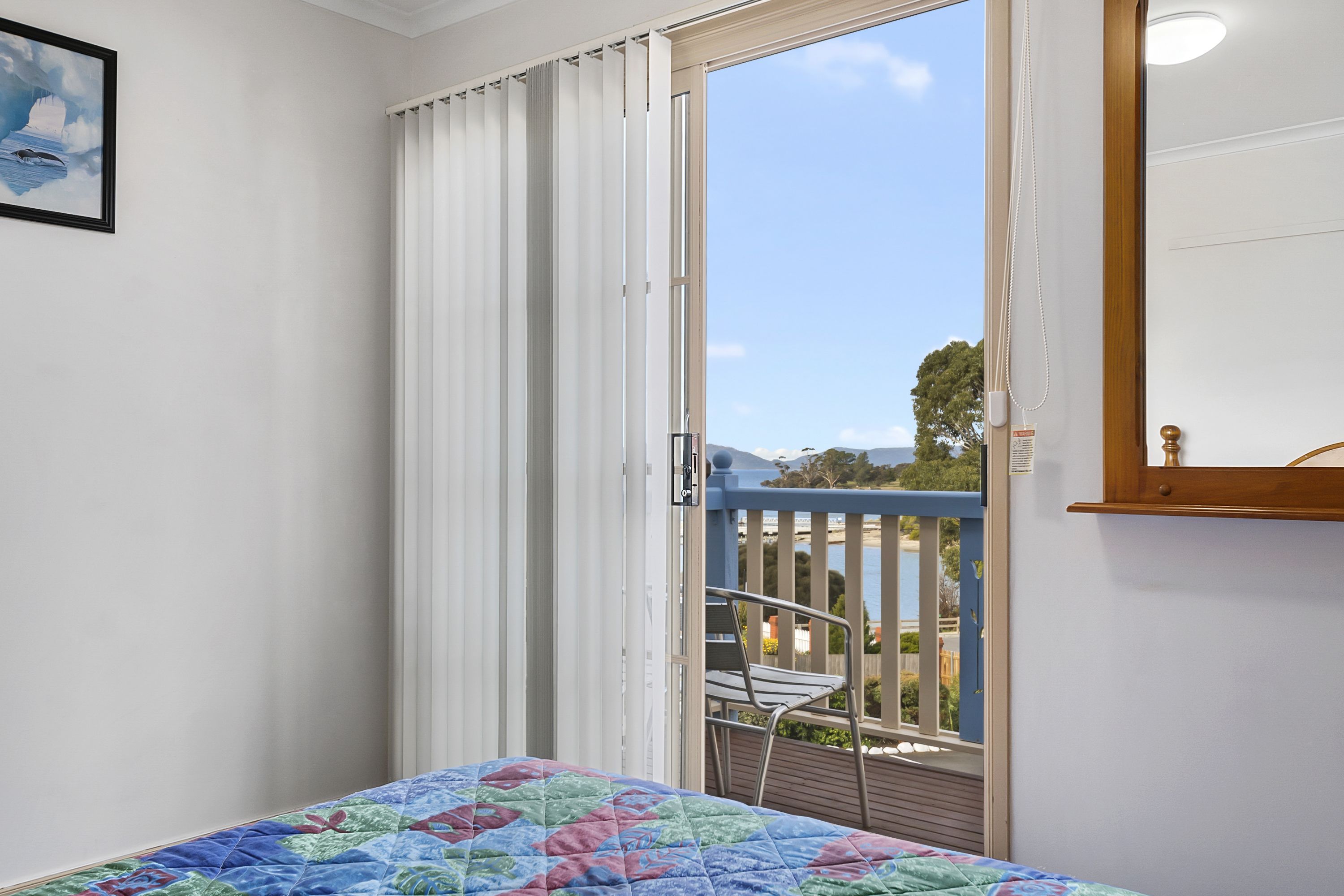 Une chambre à coucher avec un lit, une fenêtre avec des rideaux, un balcon avec une chaise et un tableau encadré au mur.