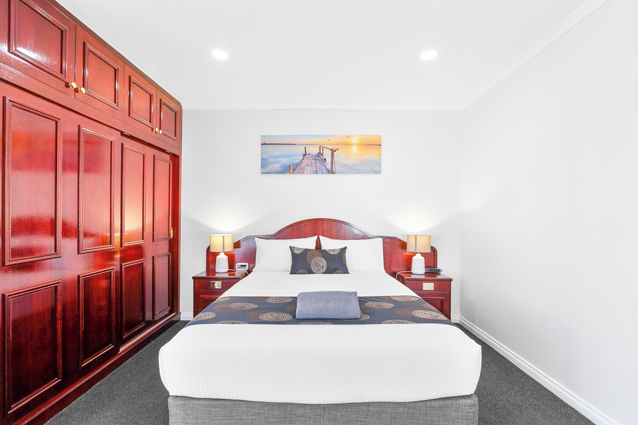 A hotel room with a large bed, wooden wardrobe, two bedside tables with lamps, and a painting of a sunset over a pier.