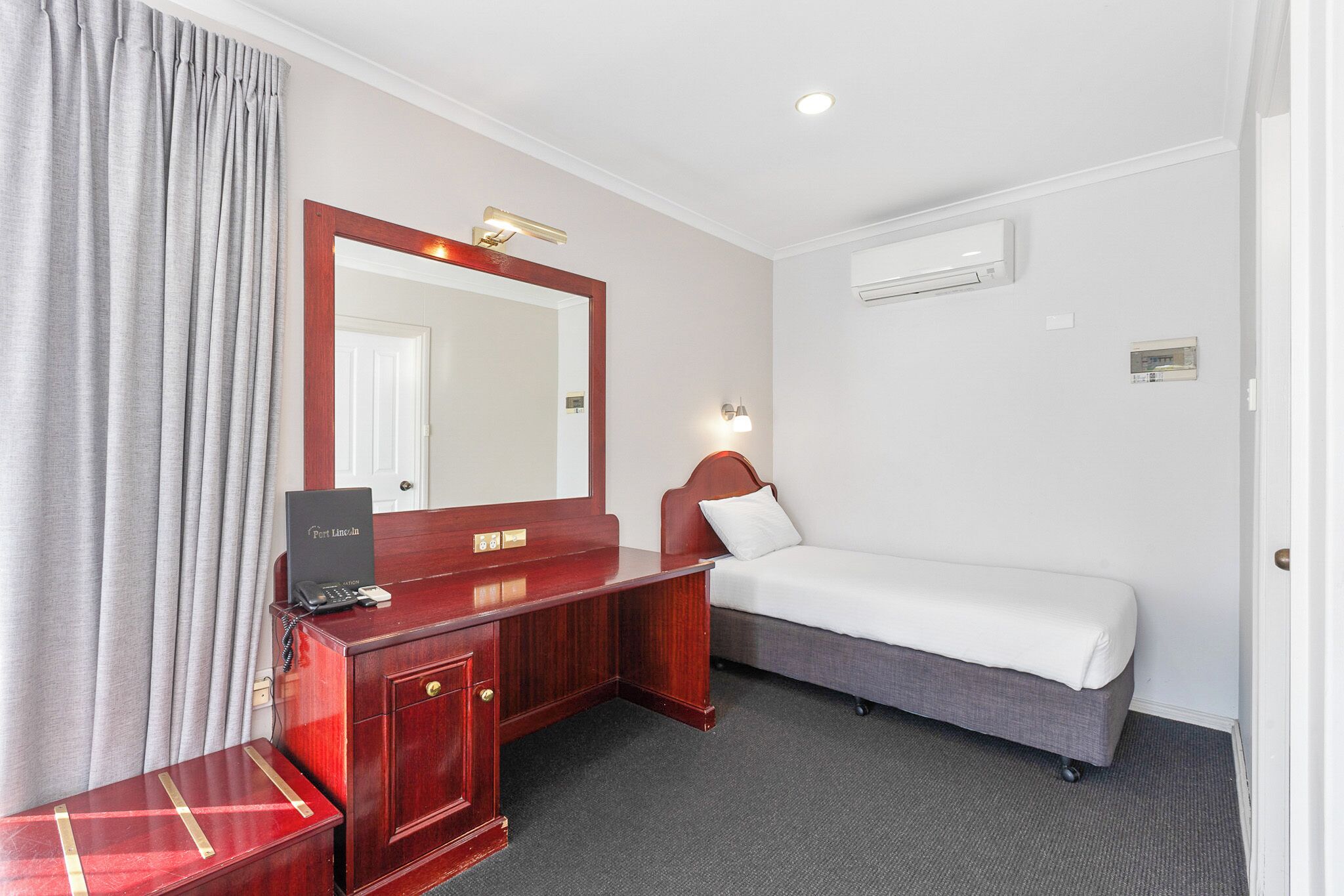 A hotel room with a wooden desk, a bed, a large mirror, and a wall-mounted air conditioner.