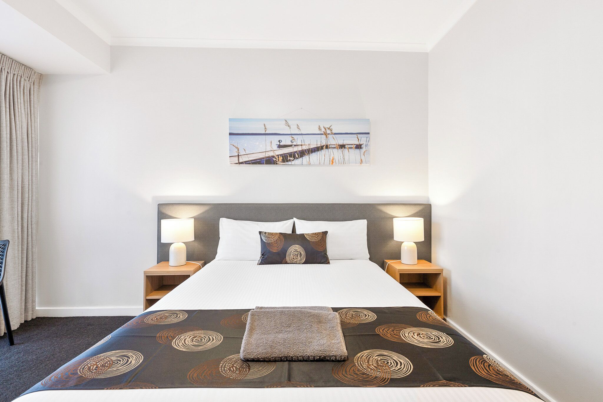 Quarto de hotel com uma cama grande, duas mesas de cabeceira com luminárias, cabeceira, quadro na parede e janela com cortinas.