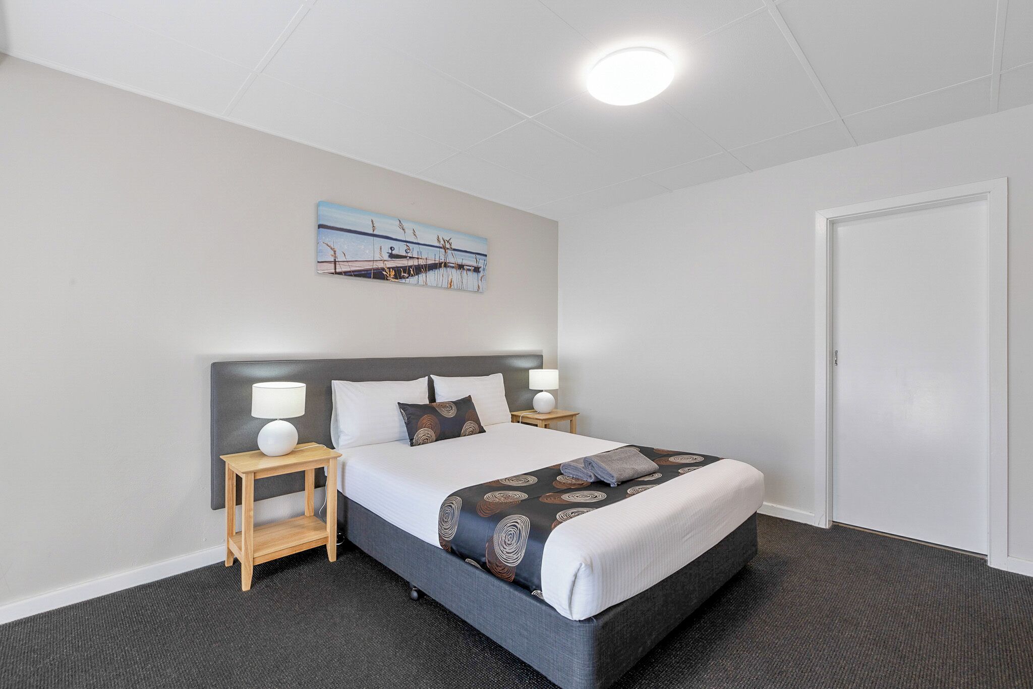 Une chambre d’hôtel avec un lit, deux tables de chevet, une commode, un tableau au mur et une porte.
