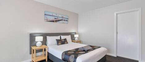 A hotel room with a bed, two bedside tables, a nightstand, a painting on the wall, and a door.