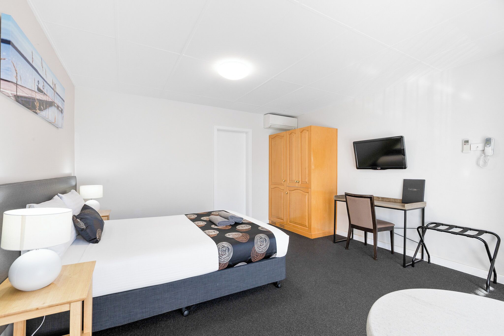 A hotel room with a bed, bedside table, lamp, desk with chair, and a TV mounted on the wall.