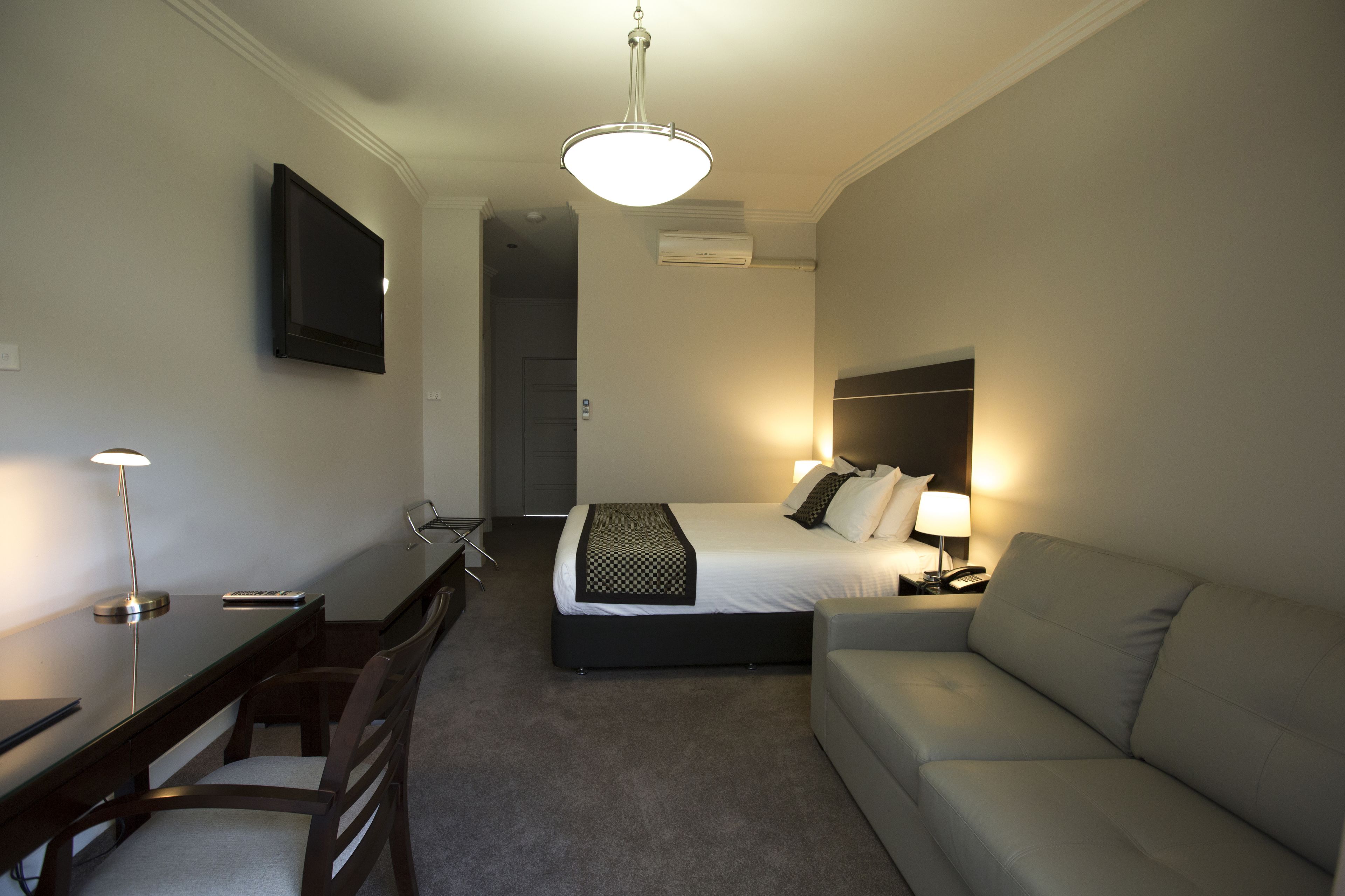 A hotel room with a bed, a desk, a chair, a sofa, a TV, and a ceiling light.