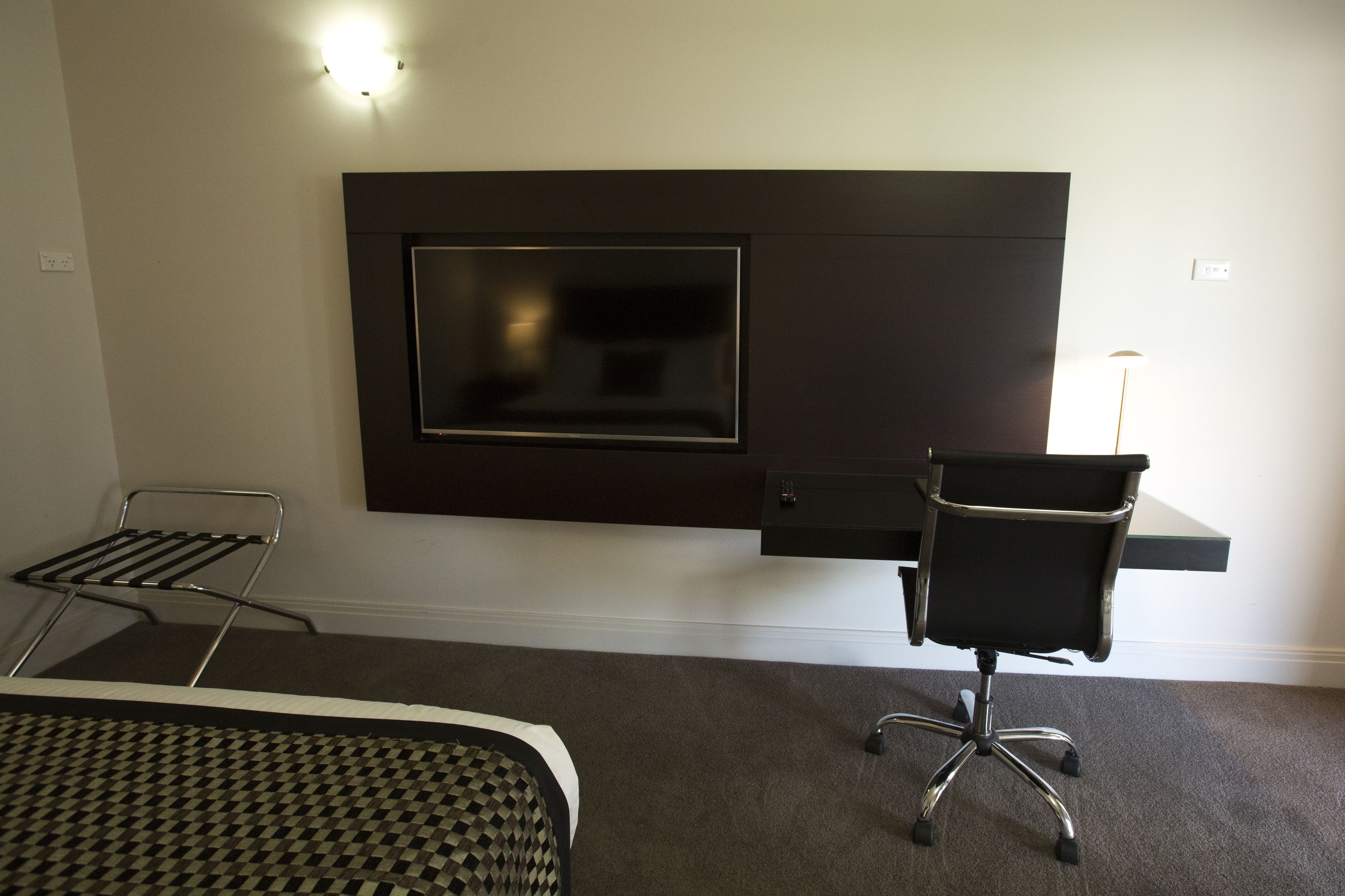 A modern hotel room with a flat-screen TV mounted on the wall, a desk with a chair, and a patterned bedspread.