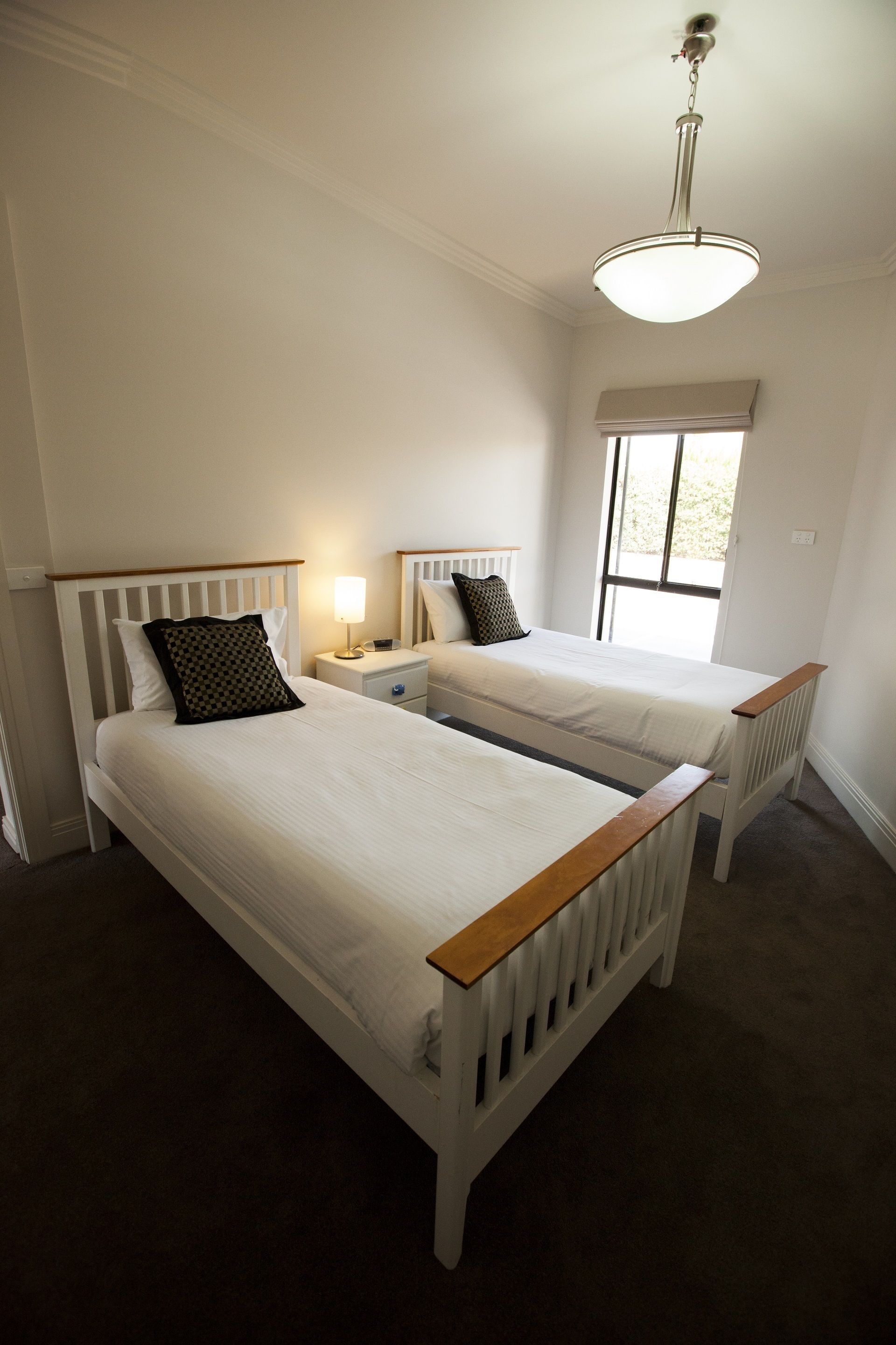 A bedroom with two single beds, a nightstand with a lamp, a window, and a ceiling light fixture.