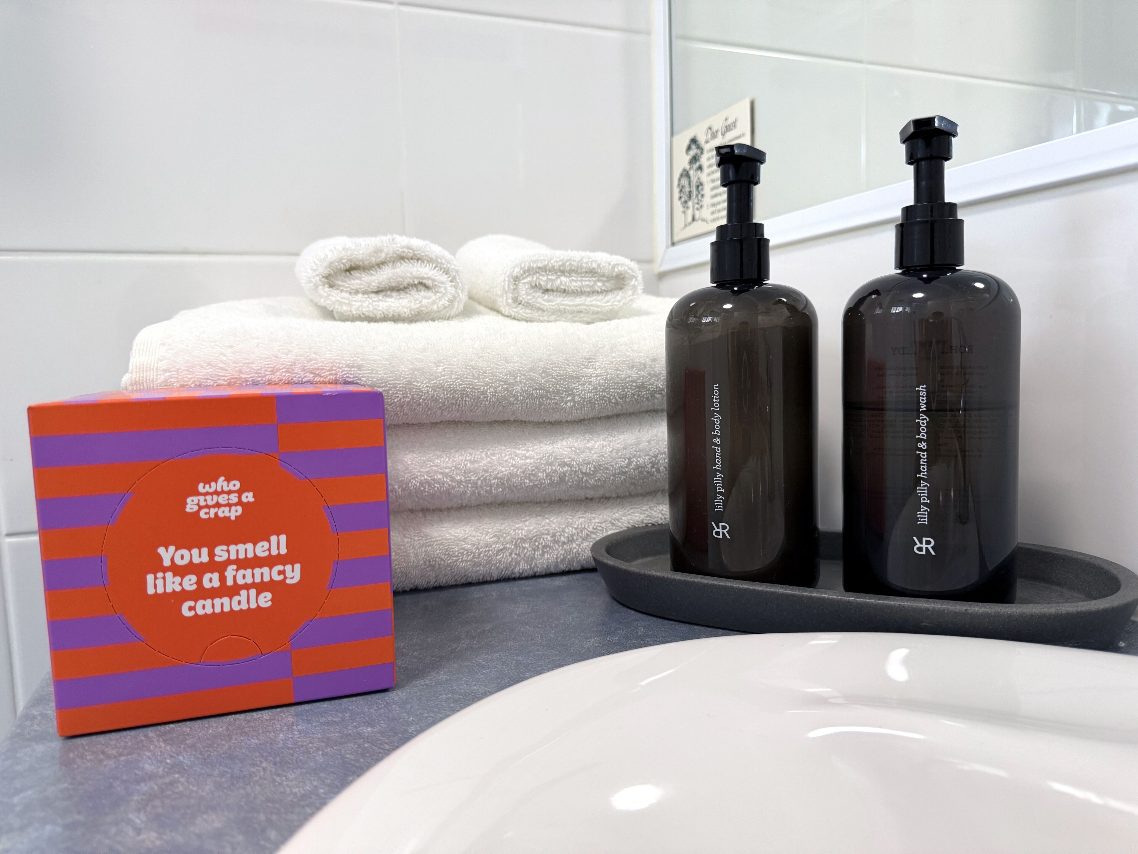 Un bagno con un lavandino bianco, due flaconi di lozione corpo, una scatola di candele profumate e asciugamani piegati con cura.