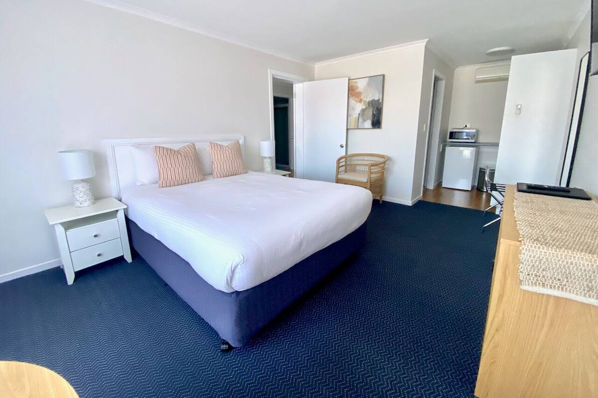 Quarto de hotel com uma cama grande, mesa de cabeceira com abajur, cadeira e escrivaninha com computador.