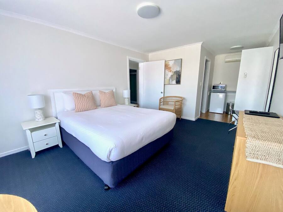 Habitación de hotel con una cama grande, una mesita de noche con lámpara, una silla y un escritorio con ordenador.