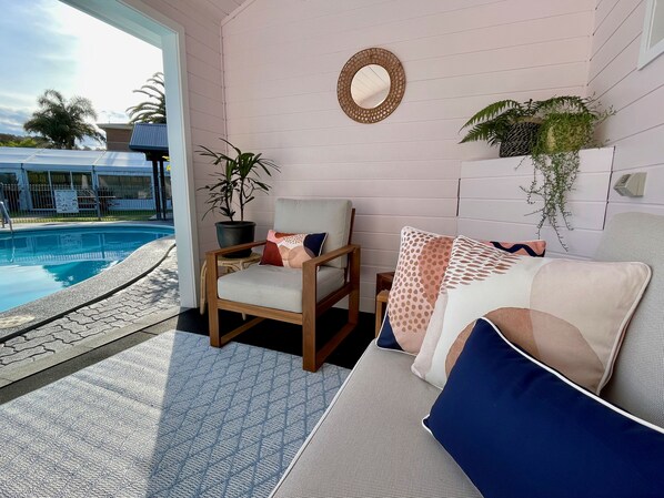 A room with a pool view, a chair with cushions, a sofa with patterned cushions, a potted plant, and a decorative mirror on the wall.