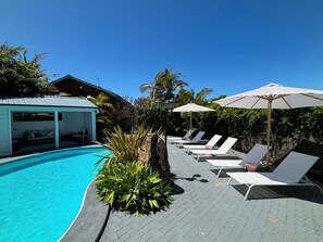 A swimming pool with lounge chairs and umbrellas, surrounded by greenery and a covered seating area. - Pelican Motor Inn (Merimbula)