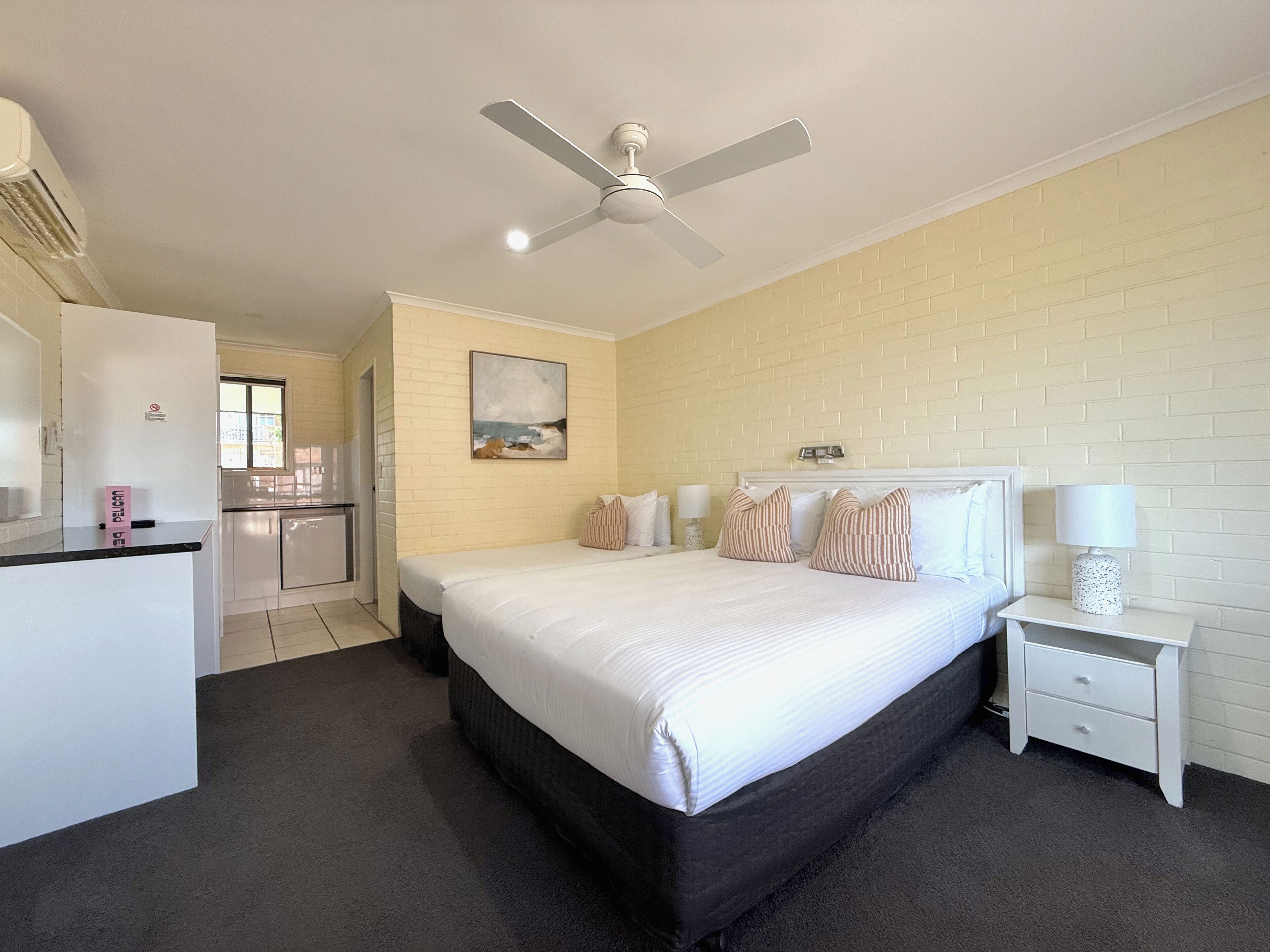 A hotel room with a bed, a nightstand, a ceiling fan, a wall-mounted painting, and a kitchenette.