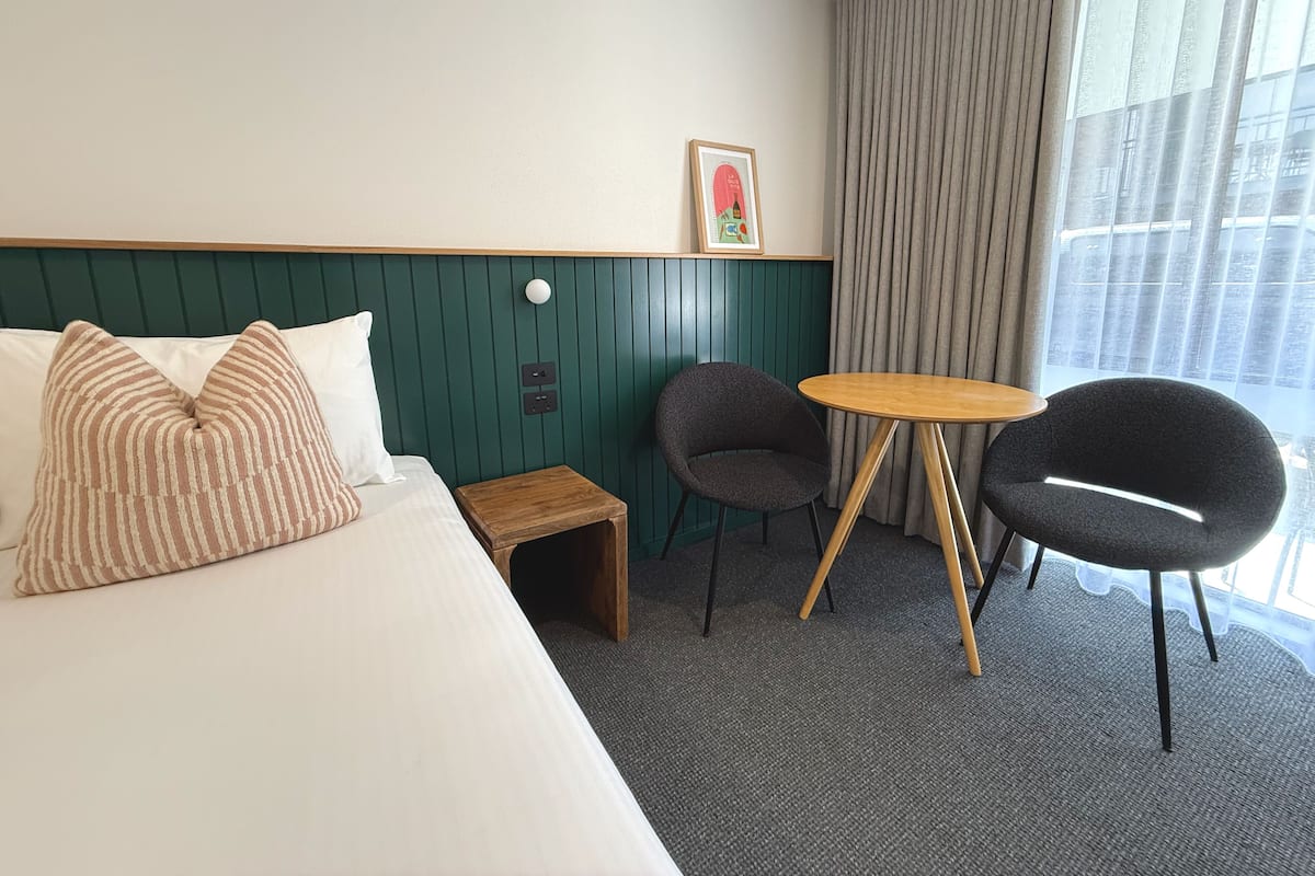 Quarto de hotel com cama, duas cadeiras, uma mesinha, um quadro na parede e uma janela com cortinas.