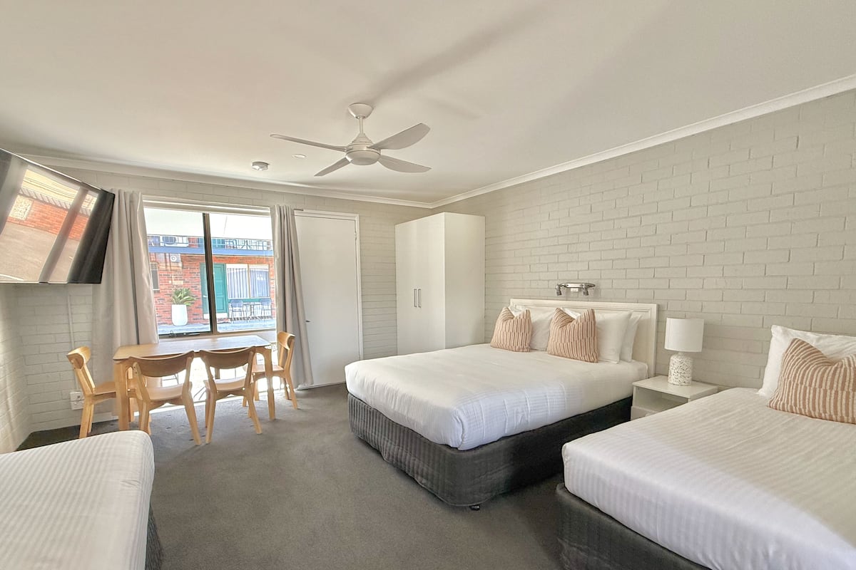 Quarto de hotel com duas camas, uma escrivaninha com cadeiras, um ventilador de teto e vista para o exterior.