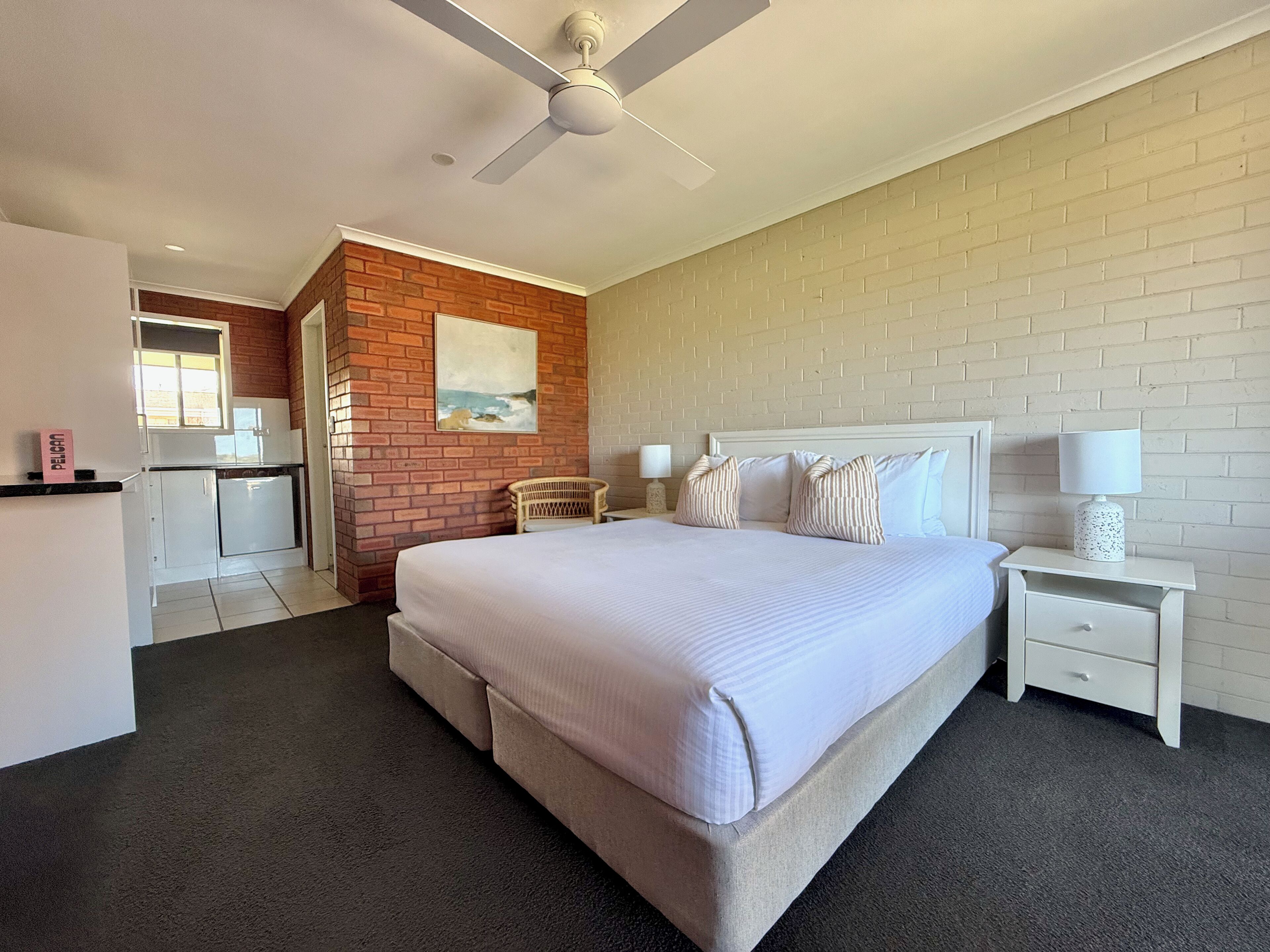 A bedroom with a large bed, a nightstand, a chair, a ceiling fan, and a brick wall.