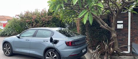 Un coche aparcado junto a un árbol, con un muro de ladrillo y una caja de servicios al fondo.