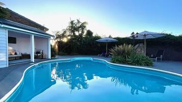 Una piscina con un área de patio techada, sillas de descanso y sombrillas.