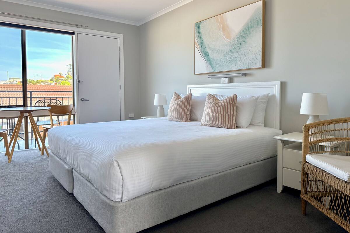 Quarto de hotel com uma cama grande, duas mesas de cabeceira com luminárias, uma cadeira de vime, uma mesa de jantar com cadeiras e uma grande janela com vista.