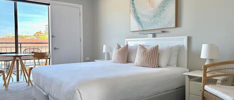 Habitación de hotel con una cama grande, dos mesitas de noche con lámparas, una silla de mimbre, una mesa de comedor con sillas y un ventanal con vistas.