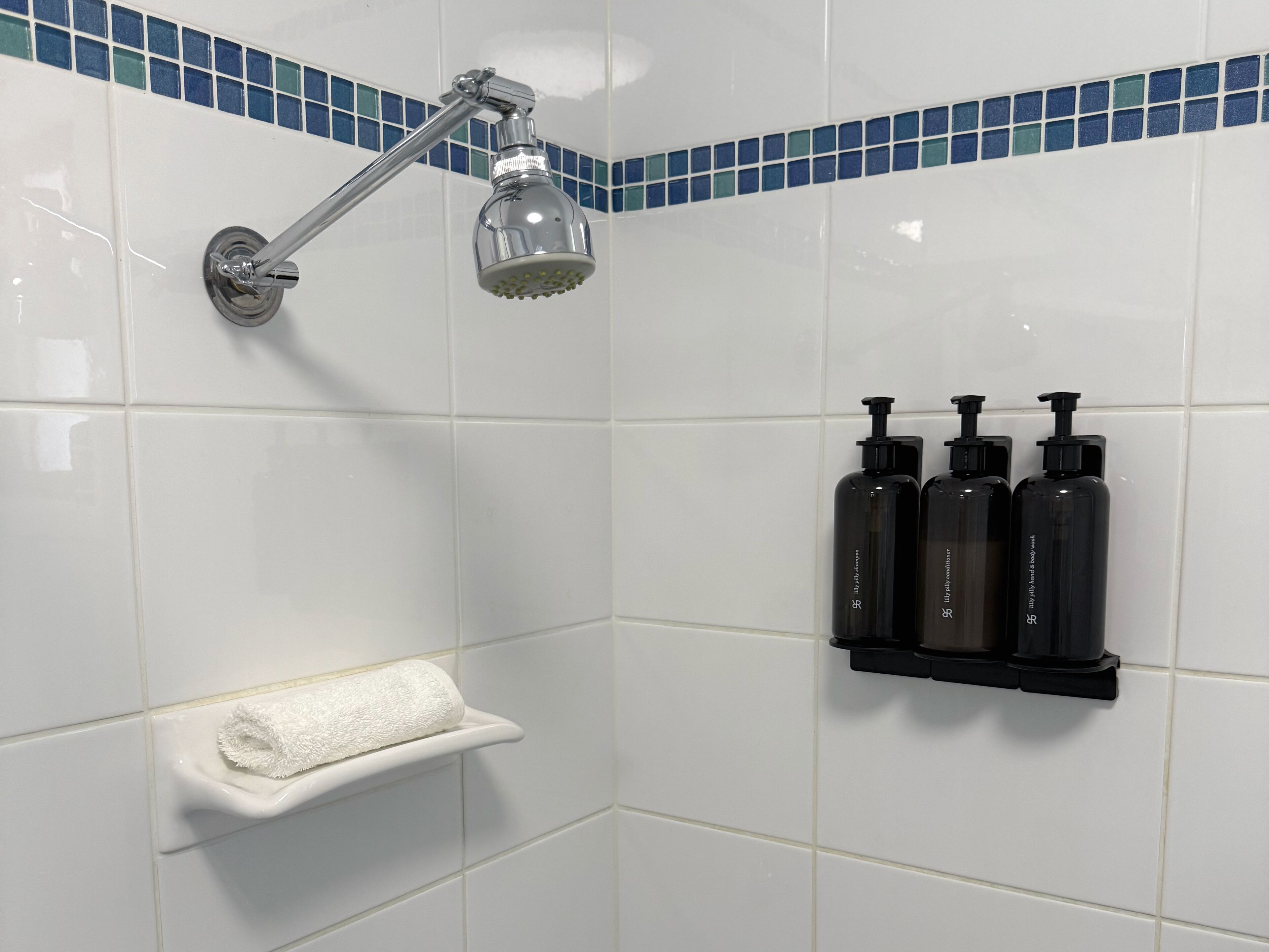Une salle de bain carrelée de carreaux blancs, une douche avec un pommeau chromé et une étagère sur laquelle sont posées trois bouteilles noires.