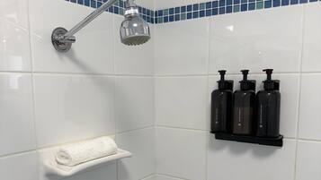 Une salle de bain carrelée de carreaux blancs, une douche avec un pommeau chromé et une étagère sur laquelle sont posées trois bouteilles noires.