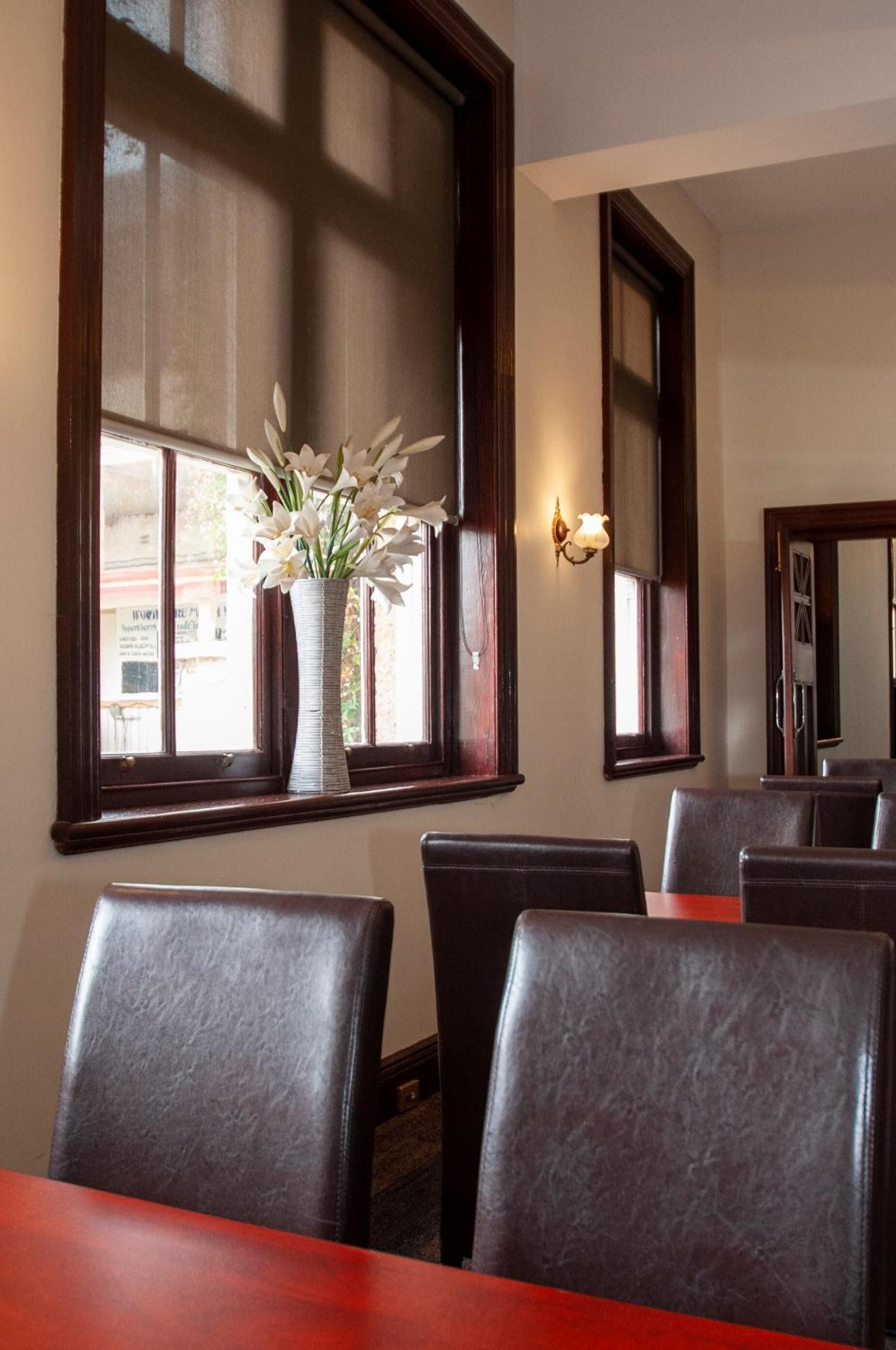 Un comedor con una mesa y sillas de madera, un jarrón con flores en el alféizar de la ventana y una lámpara de pared.