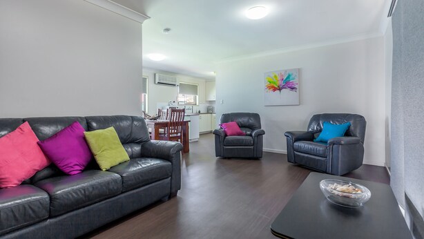 Una sala de estar con un sofá de cuero oscuro, dos sillones y una mesa de centro. Se ve en el fondo una zona de comedor con una mesa y sillas de madera.
