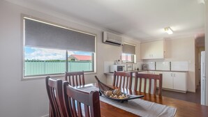 Un comedor con una mesa y sillas de madera, una cesta tejida con aperitivos, y vista a una casa a través de grandes ventanales.