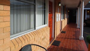 Ein überdachter Gang mit Ziegelaußenwand, großen Fenstern mit Jalousien und nummerierten Türen.