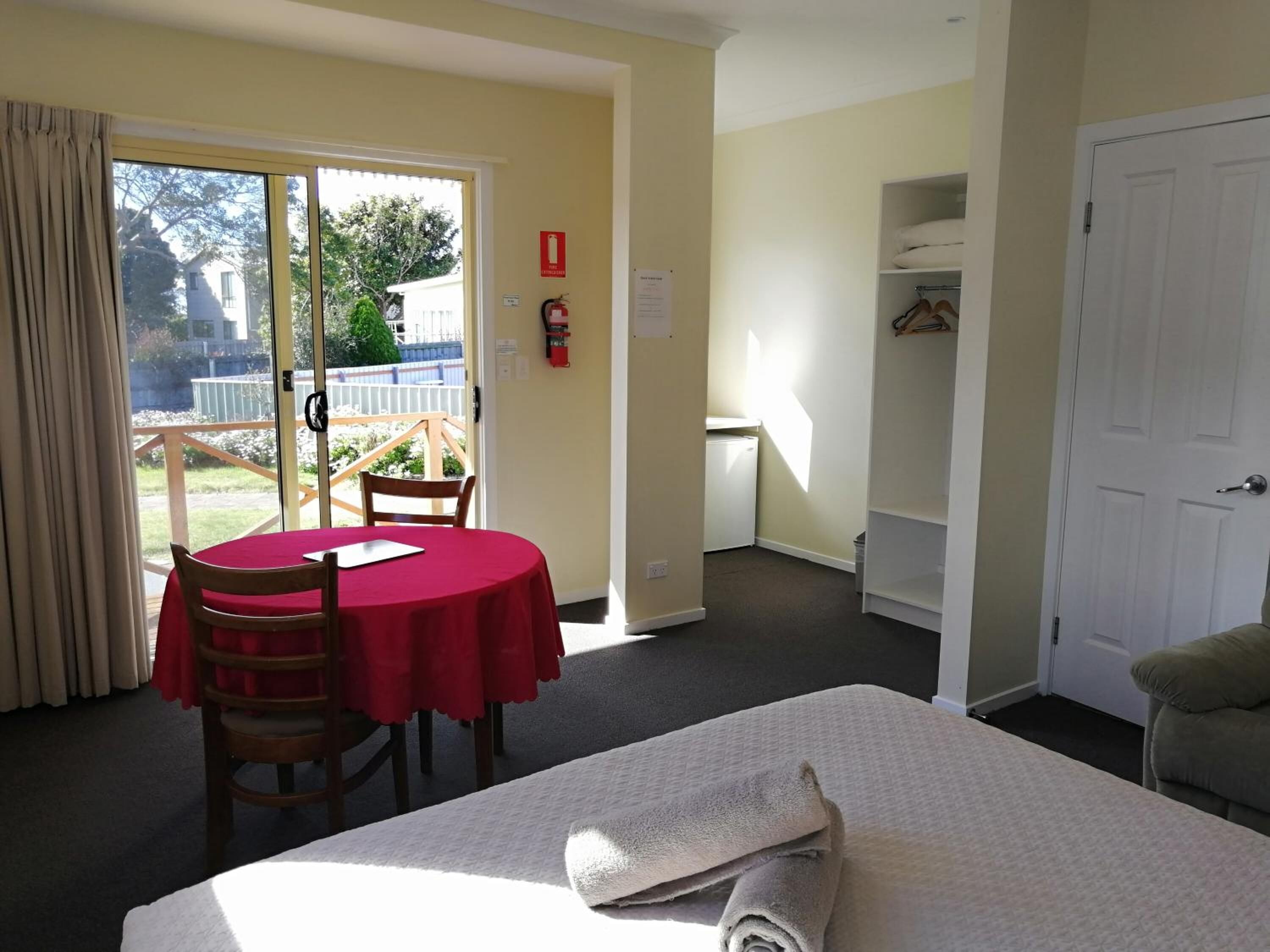 A room with a bed, a table with a red tablecloth, a chair, a wardrobe, and a door.
