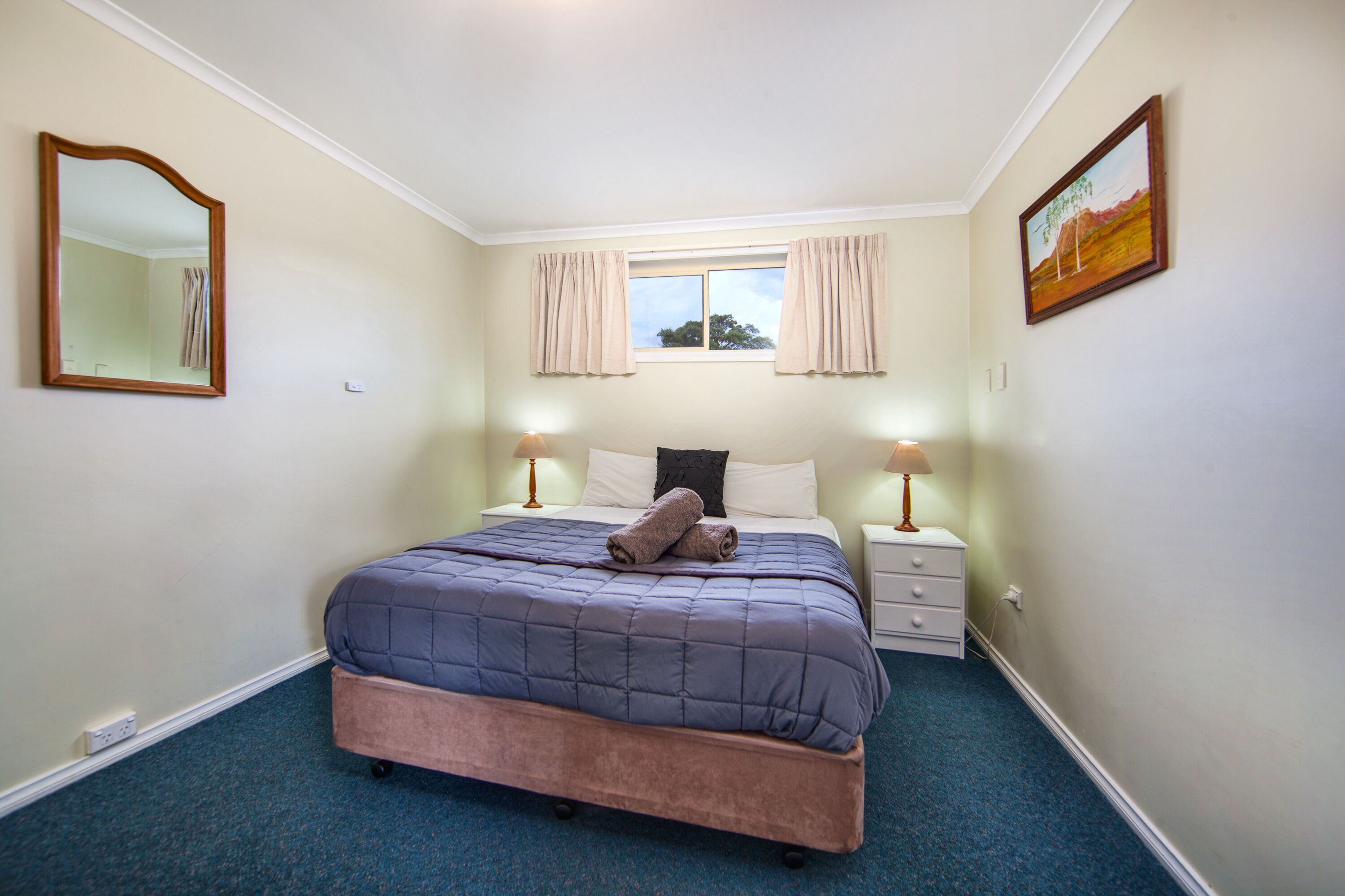 Un dormitorio con cama, lámparas de noche, una ventana con cortinas, un cuadro en la pared y un espejo.