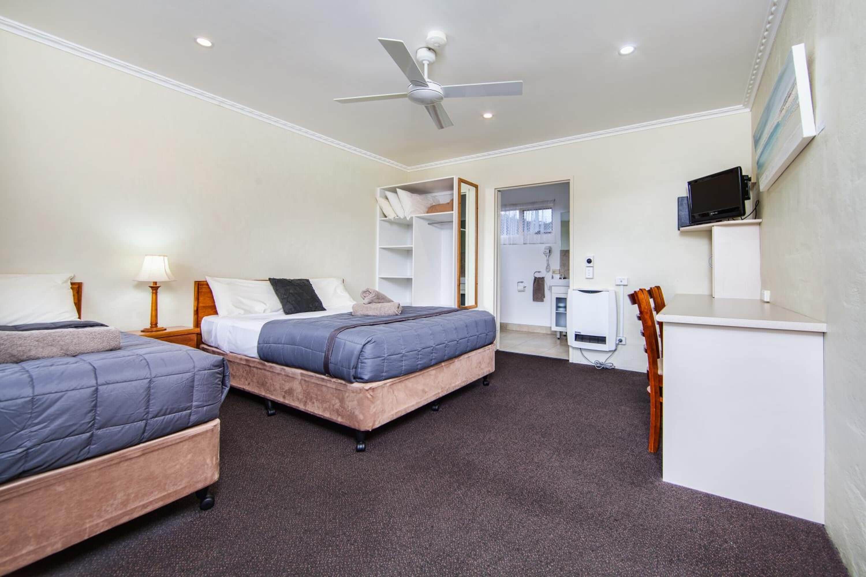 A hotel room with two beds, a desk, a chair, a TV, and a ceiling fan.