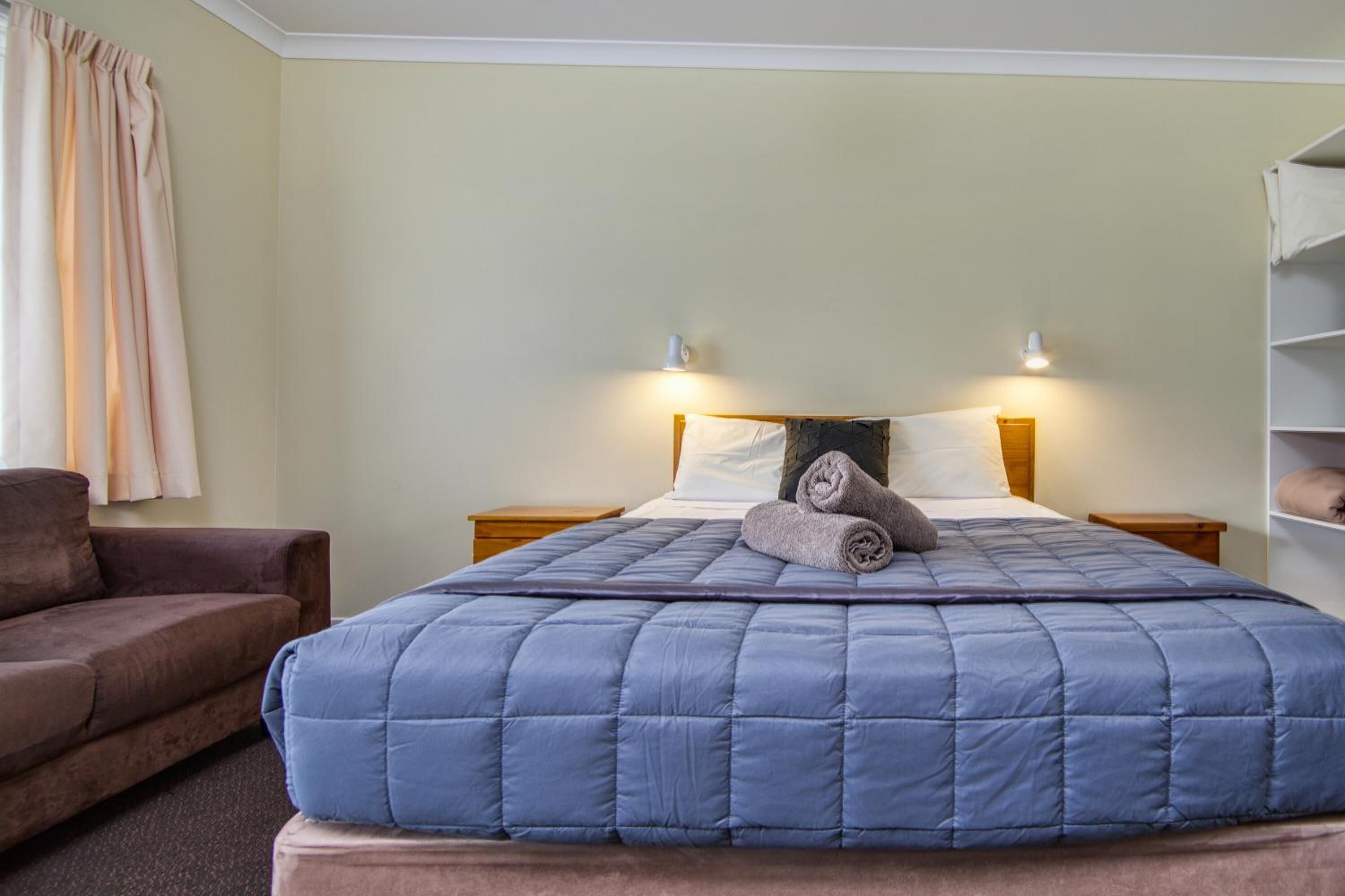 A neatly made bed with a blue quilt, two bedside lamps, and a brown couch in a room with light-colored walls.