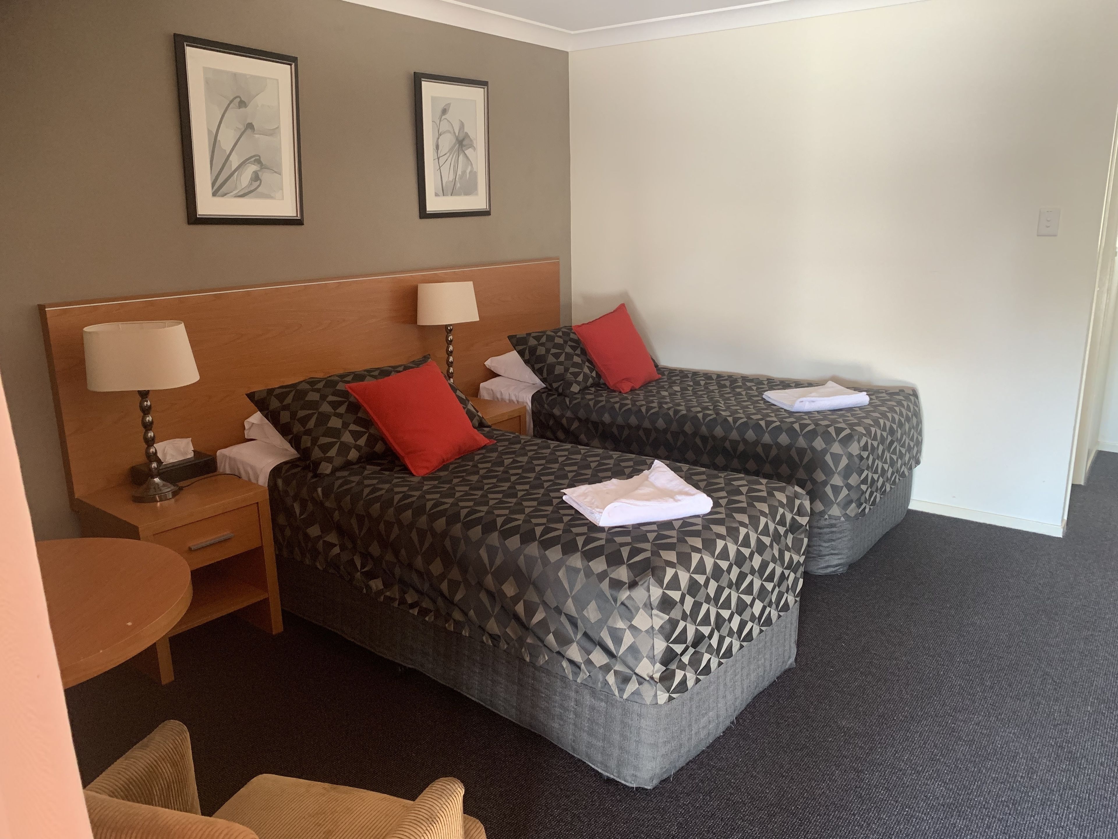 Habitación de hotel con dos camas, cada una con una colcha estampada y almohadas rojas. Hay mesitas de noche con lámparas y una pequeña mesa con una silla.