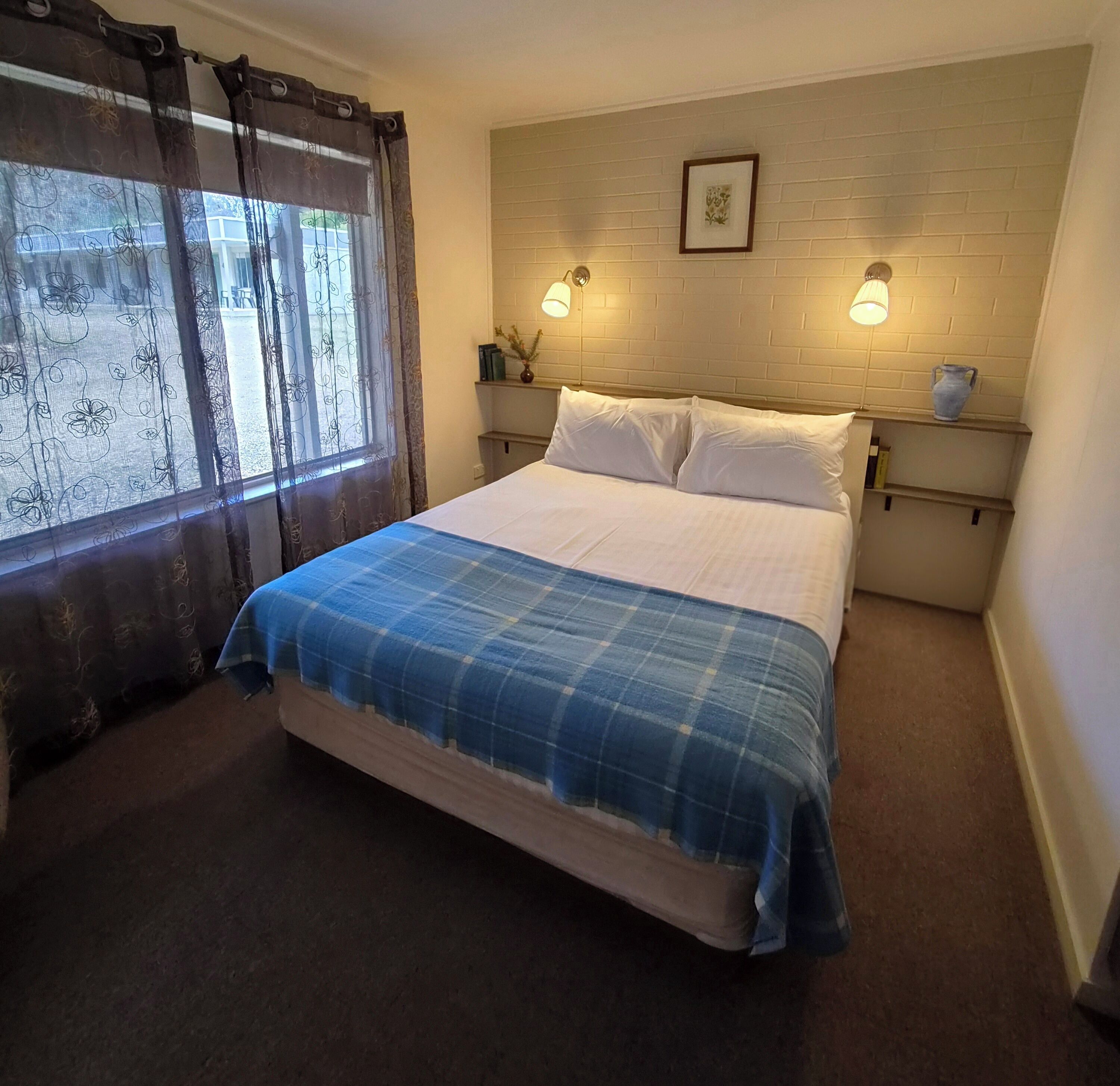 A bedroom with a bed, two bedside tables, a window with curtains, a wall-mounted picture, and a brick wall.