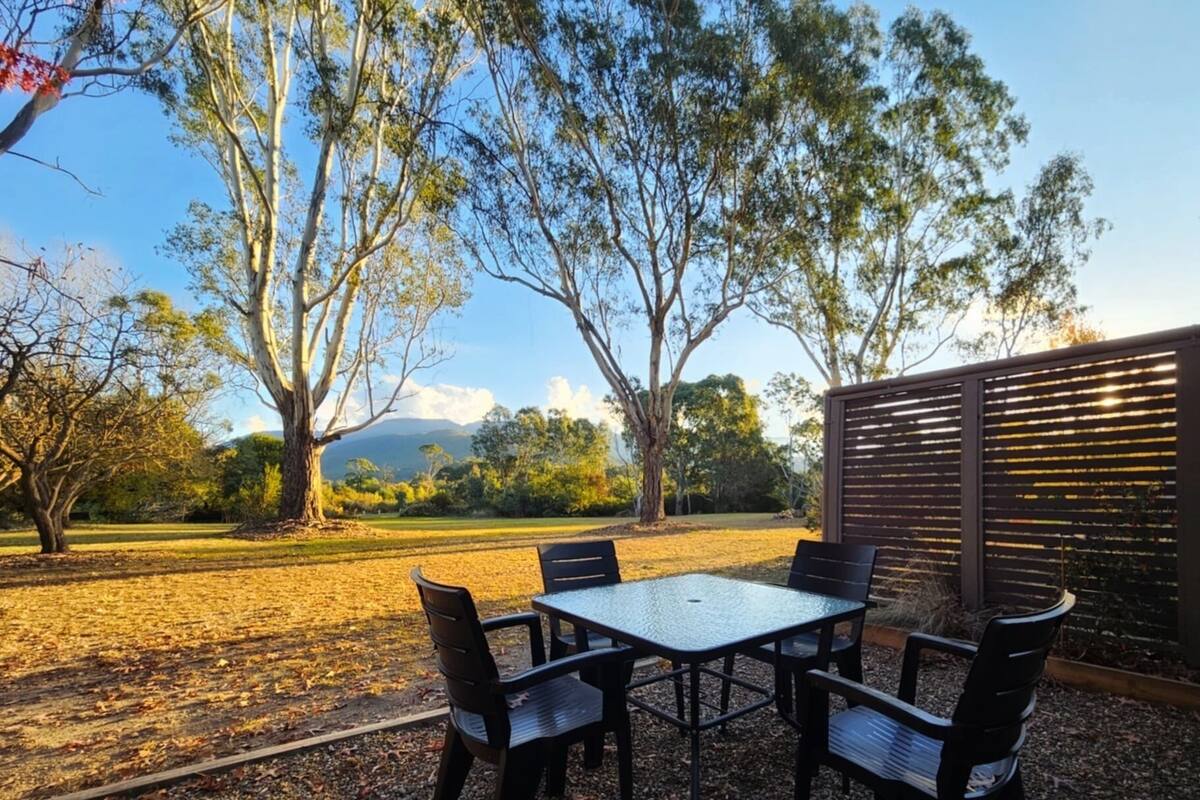 Un espacio al aire libre con mesa y sillas, rodeado de árboles y vistas a un paisaje lejano.