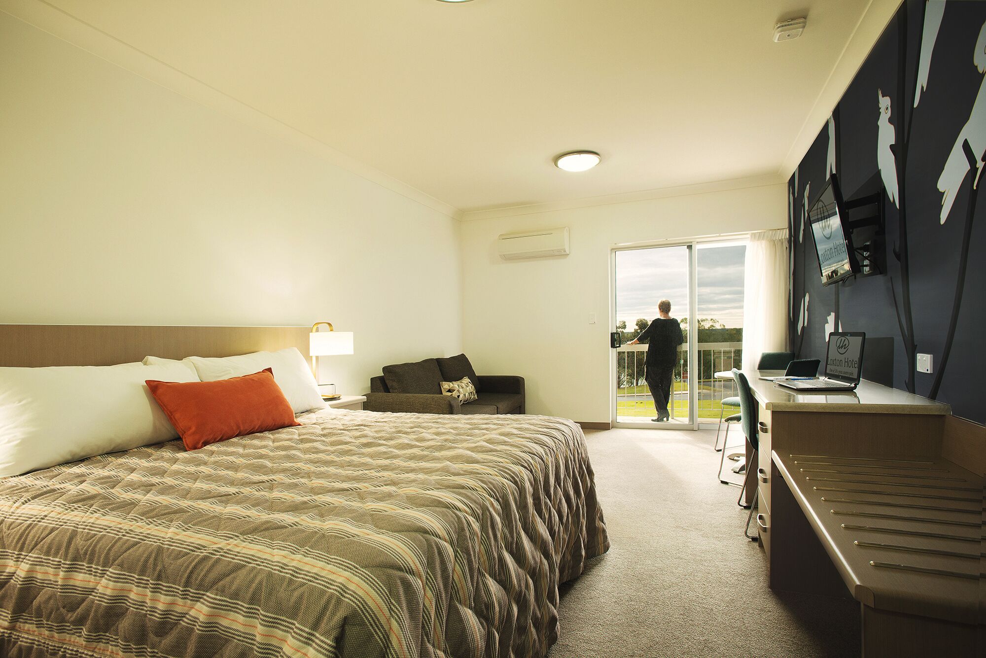 A hotel room with a bed, a desk, a chair, a sofa, and a sliding door leading to a balcony.
