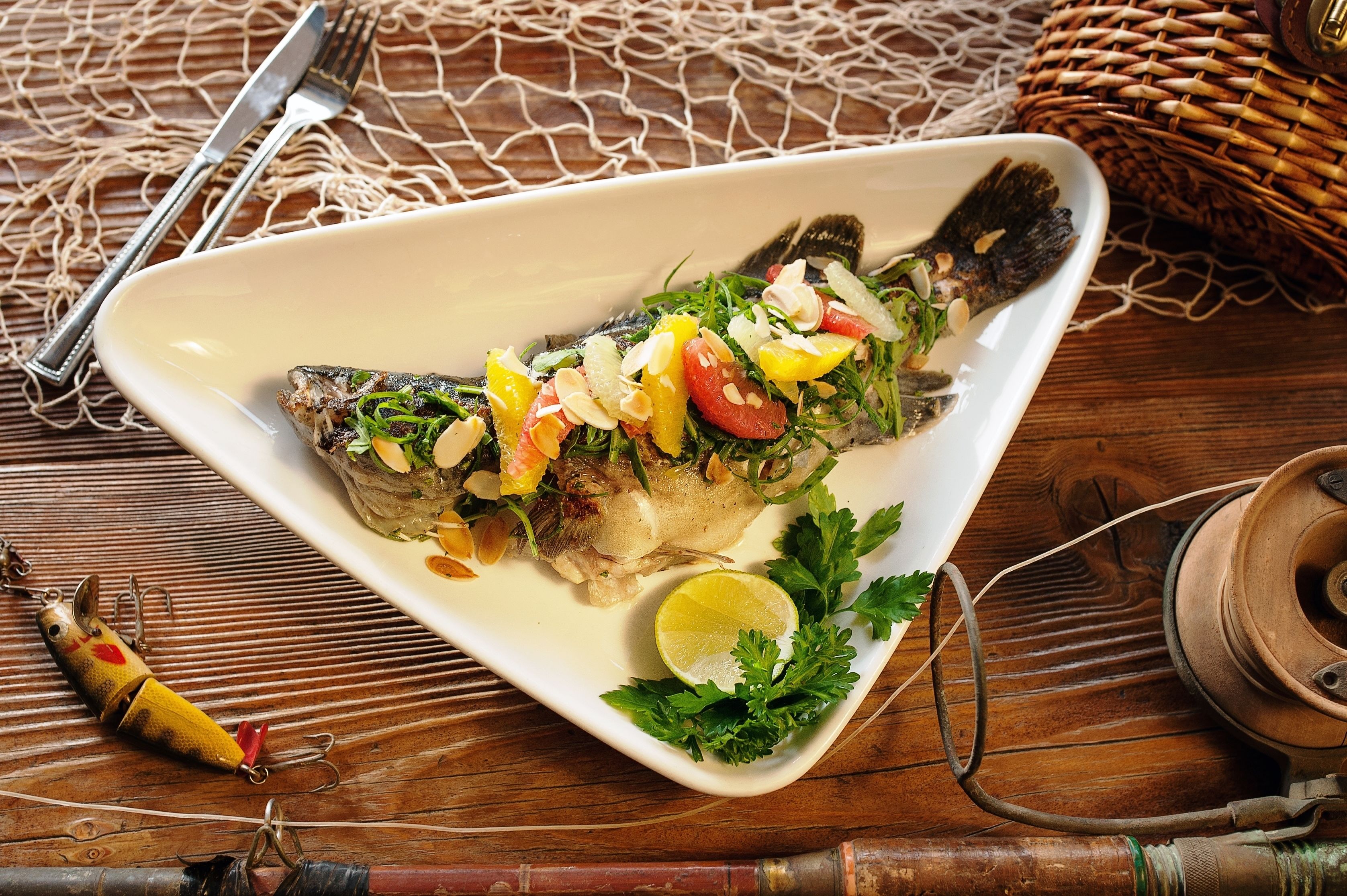 Ein weißer Teller mit gegrilltem Fisch, belegt mit Gemüse und Kräutern, dazu eine Zitronenspalte und Petersilie.