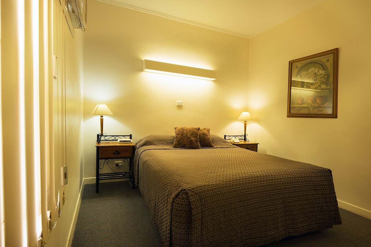 A hotel room with a bed, two bedside tables, two lamps, a painting on the wall, and a carpeted floor.