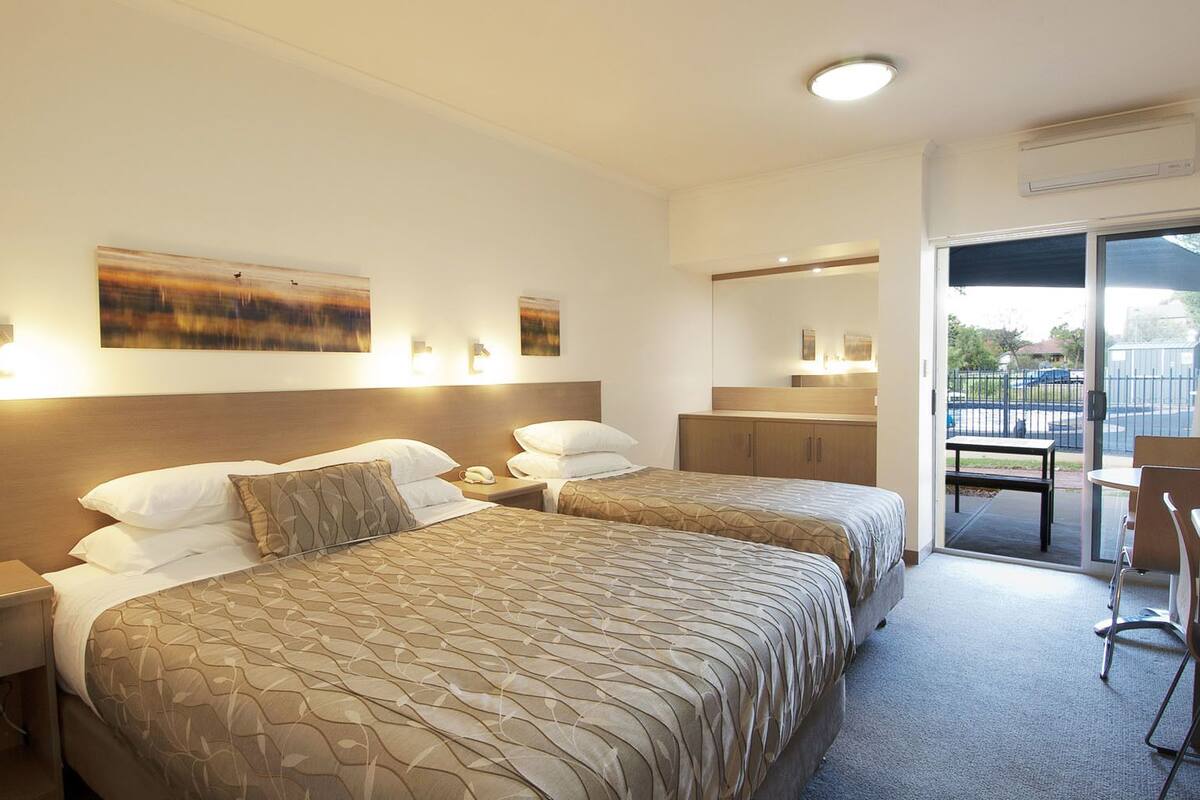 A hotel room with two beds, a small table, a chair, and a balcony with a view of the outside.