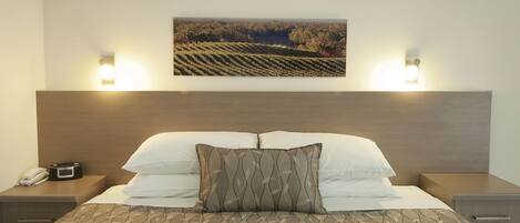 A hotel room with a bed, two bedside tables, a nightstand with a phone, and a wall-mounted picture of a vineyard.