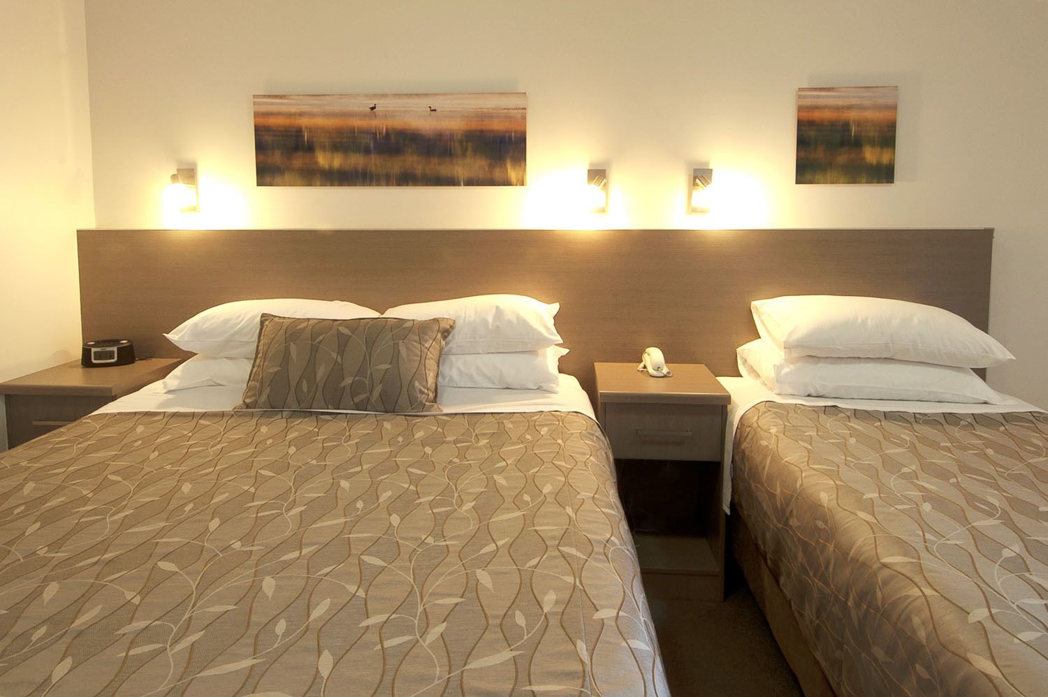 A hotel room with two beds, each with a patterned bedspread and white pillows. There are bedside tables with lamps and a phone on one side.
