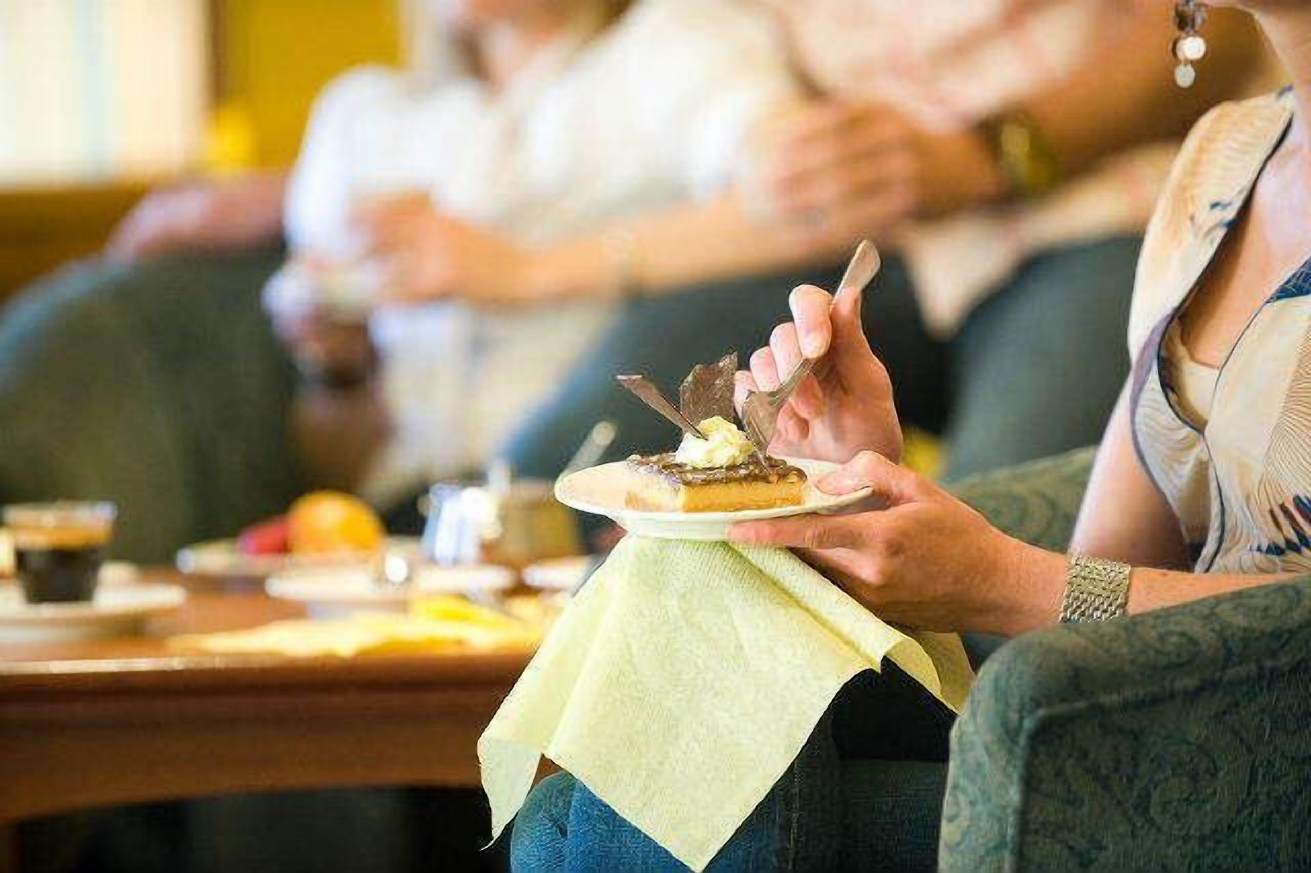 Eine Person hält einen Teller mit Dessert und sitzt an einem Tisch mit gelber Tischdecke.