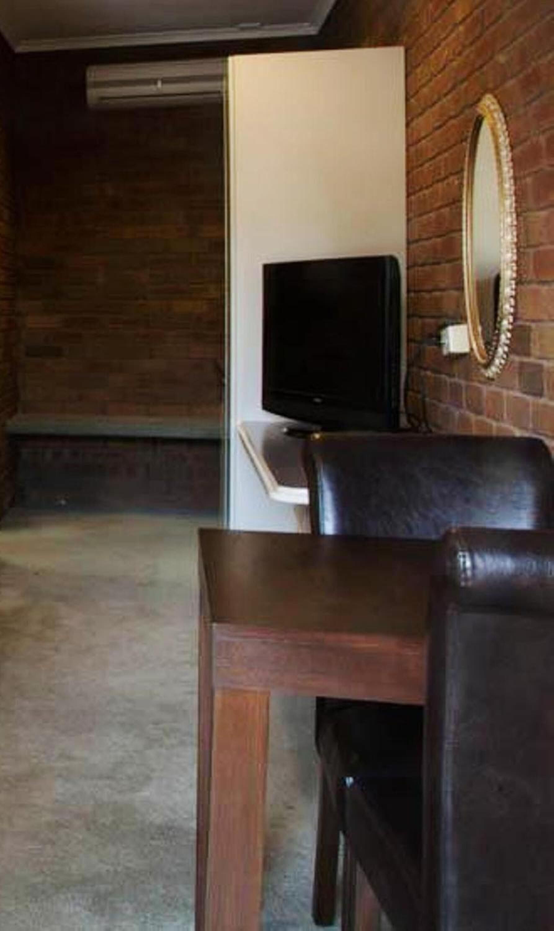 A room with a brick wall, a flat-screen TV on a white stand, a wooden table, and a leather chair.