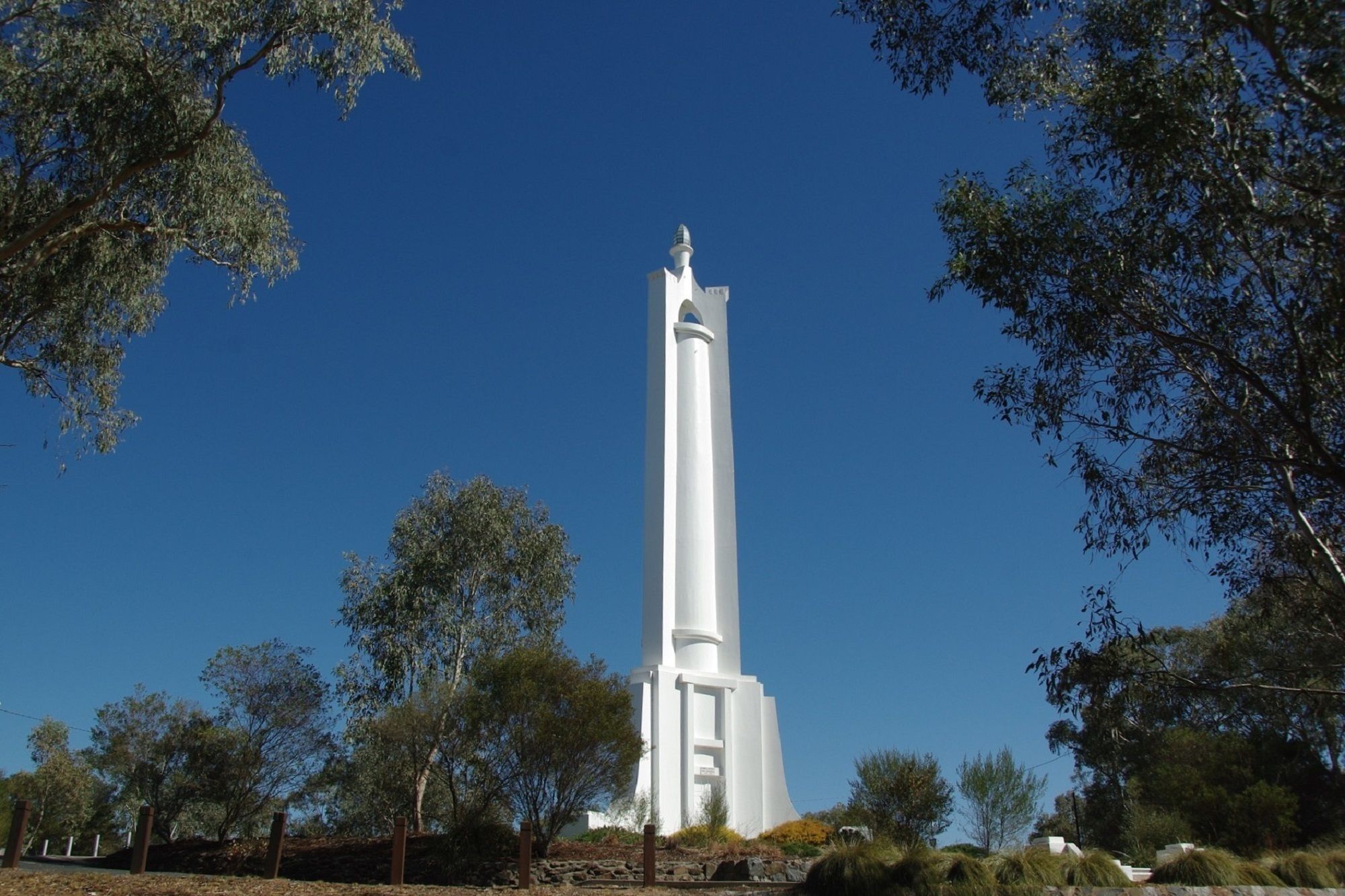 Una alta torre blanca con un pico apuntado, rodeada de árboles y un cielo azul despejado.