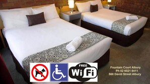 A hotel room with two beds, each with a patterned bedspread and a rolled towel. There are nightstands with lamps, a brick wall, and a sign indicating no dogs and free Wi-Fi. - Fountain Court Motor Inn Albury (Albury)